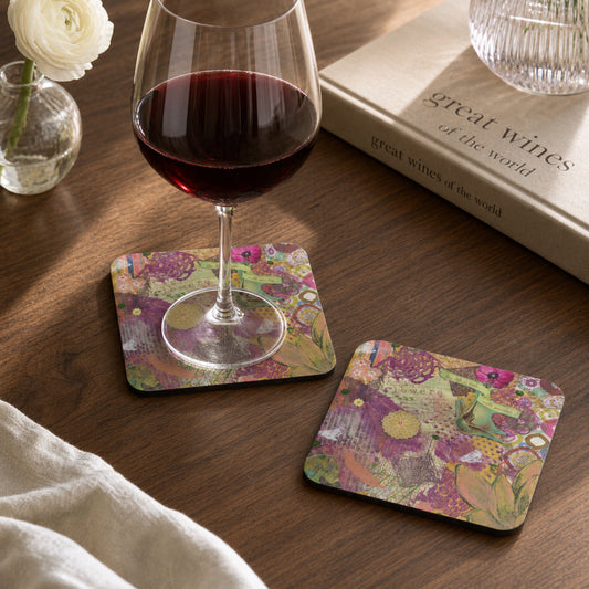 Two square art coasters with a colorful floral and botanical patchwork pattern are shown on a wooden table, one holding a glass of red wine and the other empty. A book, a small vase with a white rose, and a folded cream cloth are also visible in the scene.