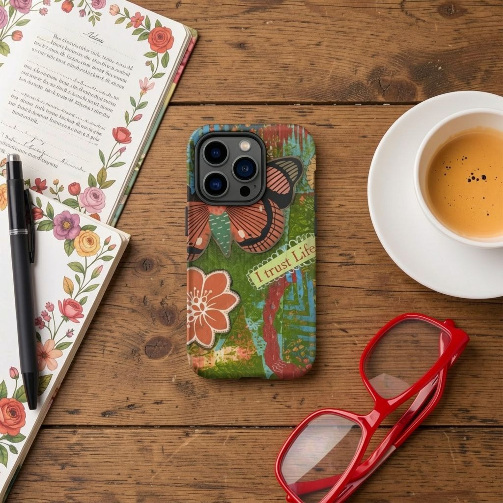 The Artistic Butterfly phone case for iPhones on a wooden table with some stationary, a pen, red framed eyeglasses and a cup of espresso.