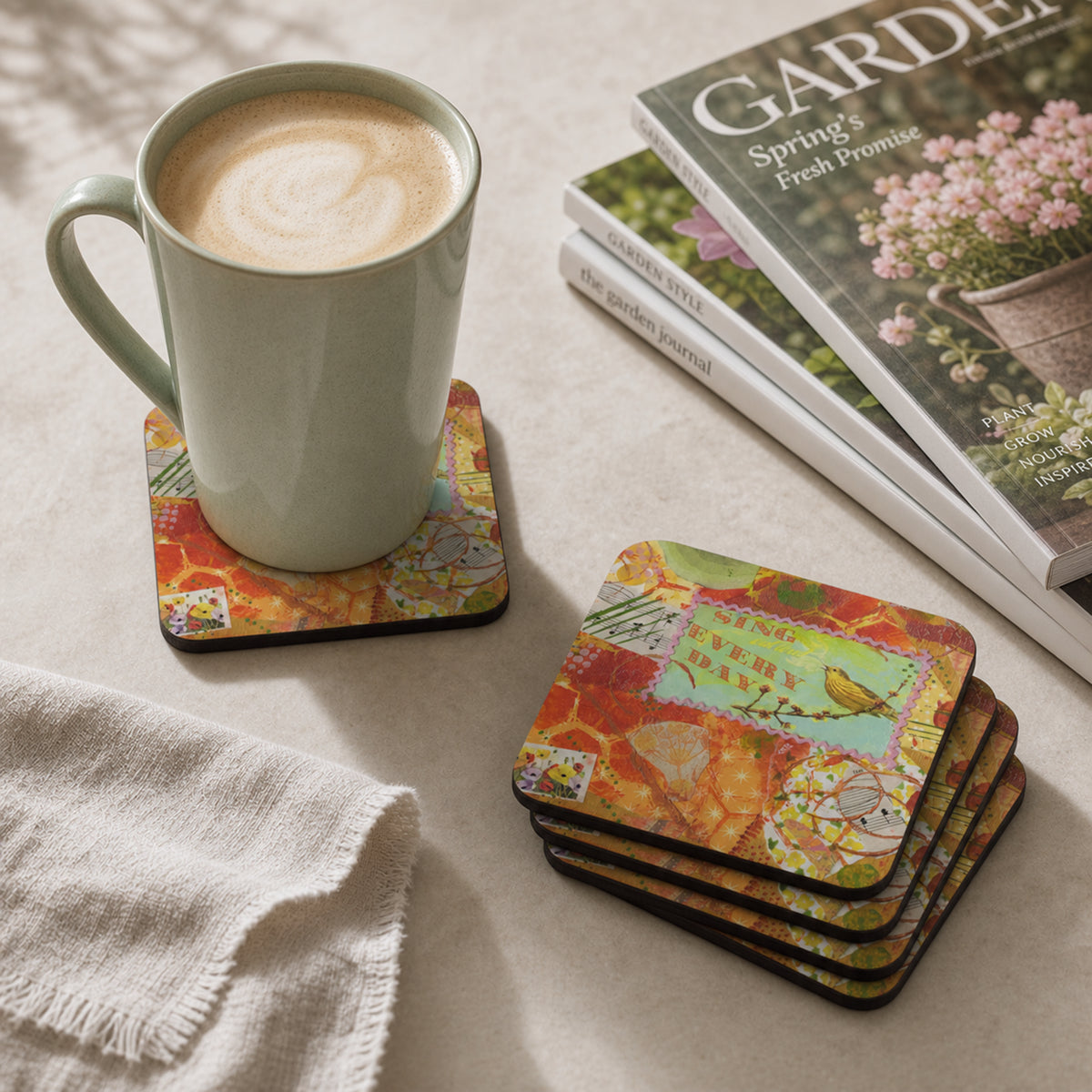 A set of square coasters with a colorful layered design with a bird on it, shown in a styled tabletop scene. One coaster holds a light green ceramic mug filled with latte art coffee, while a stack of four coasters sits beside it on a beige fabric surface next to a folded white cloth and a magazine.
