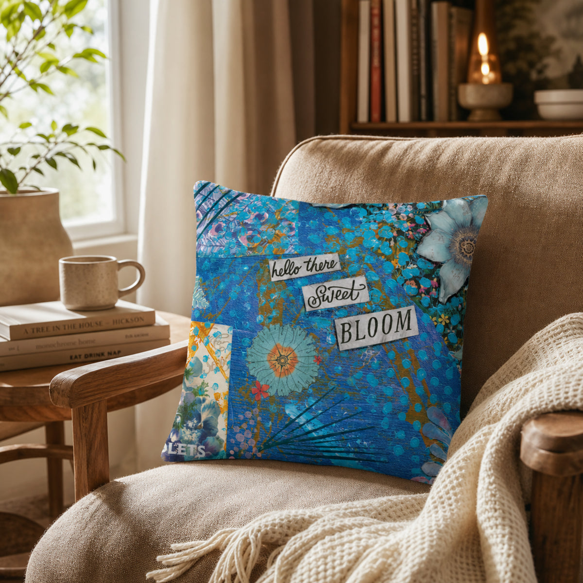 A decorative square throw pillow with a blue floral pattern and the words 'hello there Sweet BLOOM' sits on a beige armchair in a cozy living room setting.