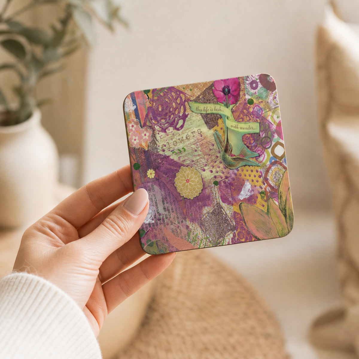 A hand holding a square coaster featuring a colorful, layered-style collage pattern with floral elements, postcards, and green ribbons in shades of purple, pink, yellow, and green.