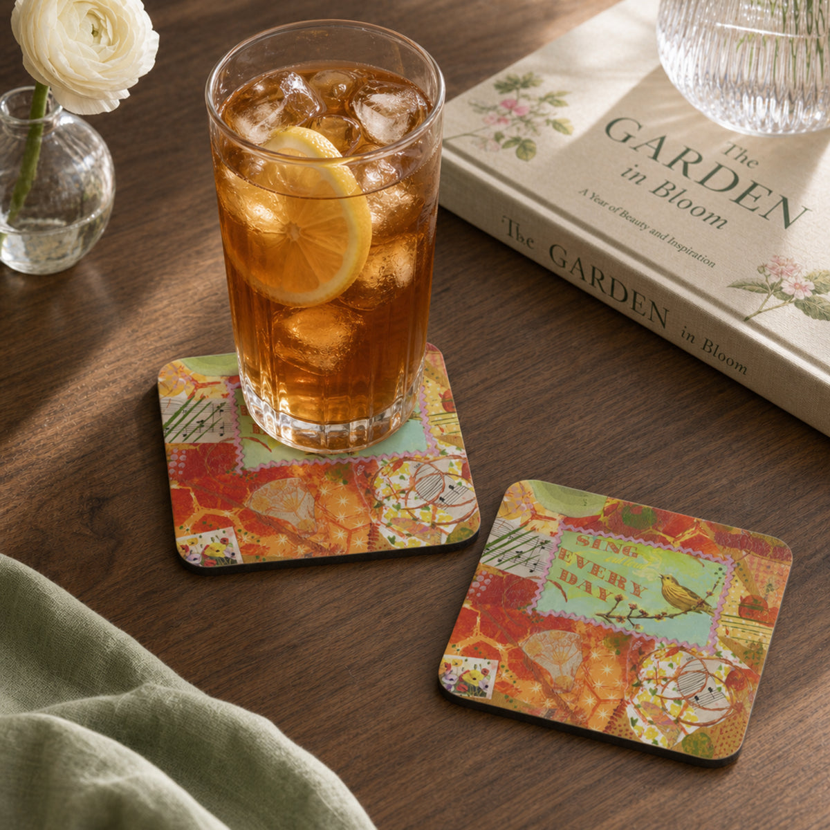 Two square coasters with a colorful layered art design are shown on a wooden table. One coaster holds a glass of iced tea with a lemon slice, and the other lies beside it.