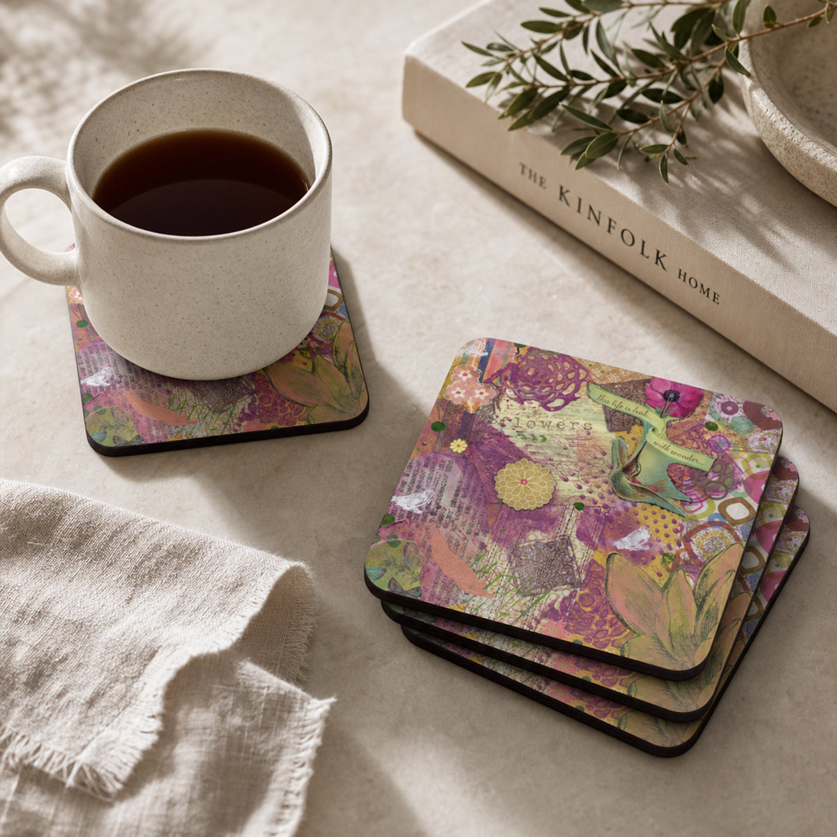 A set of square coasters with a colorful floral and botanical collage pattern, shown on a light beige tabletop. One coaster holds a white speckled ceramic mug filled with dark coffee, while a stack of coasters sits nearby.