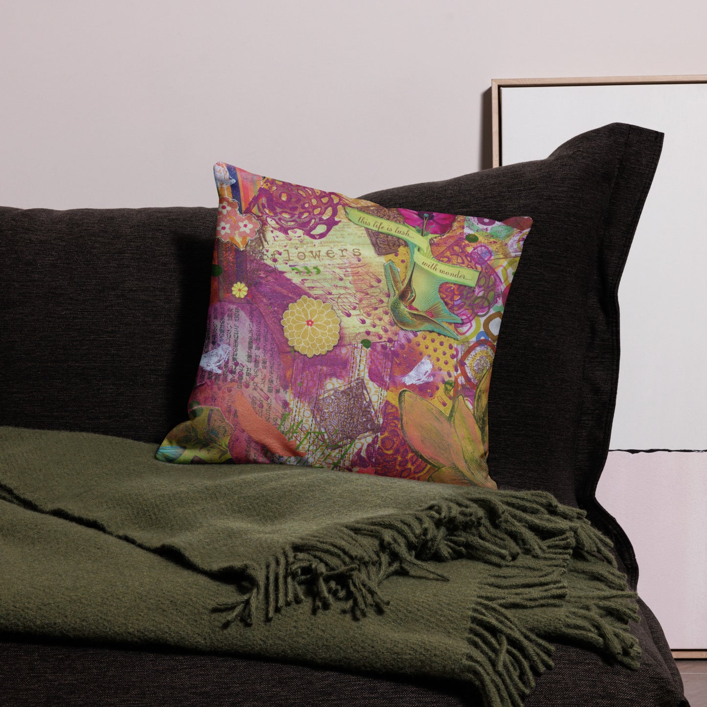 A square throw pillow with a vibrant, multicolored abstract and floral pattern sits on a dark gray sofa, styled with an olive green fringed throw blanket.
