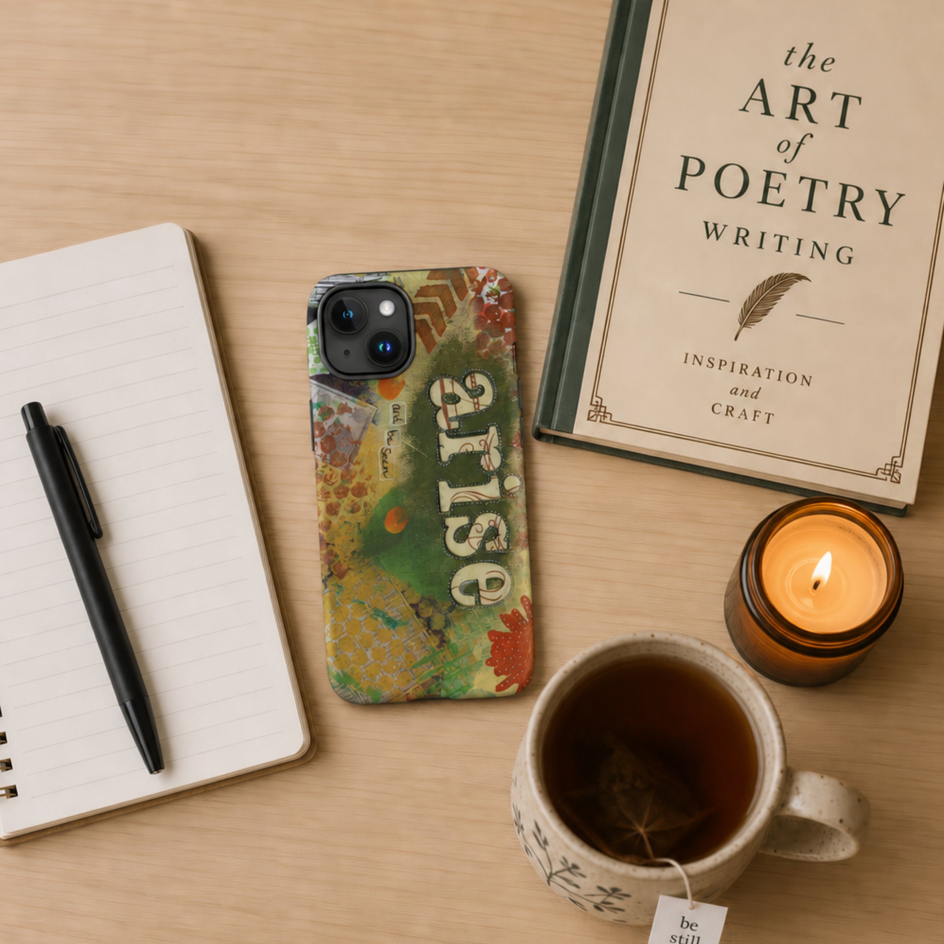 Phone with 'arise' case, notebook, pen, mug of tea, and candle on a wooden surface.