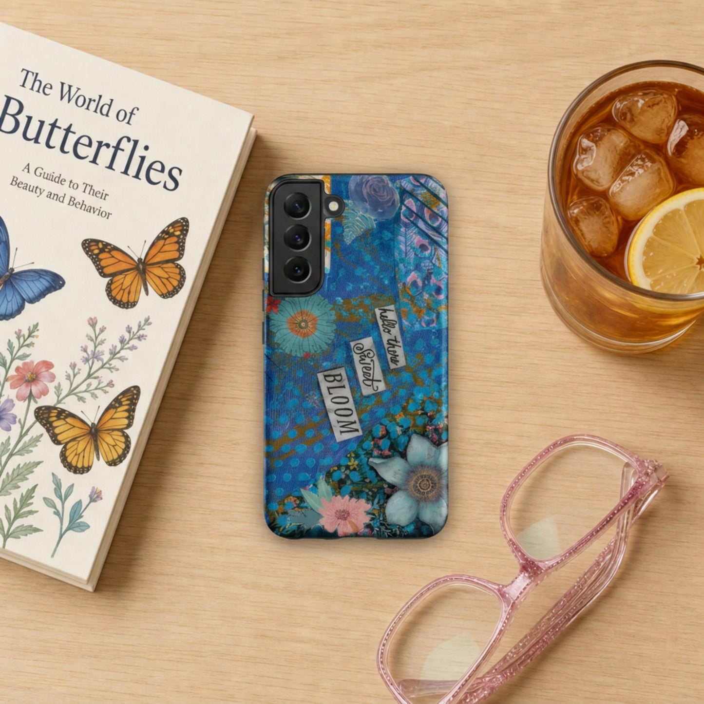 image of the blue and pink floral Samsung case with peacock feather layers and a quote, laying on a wooden table with book, eyeglasses and iced tea nearby.