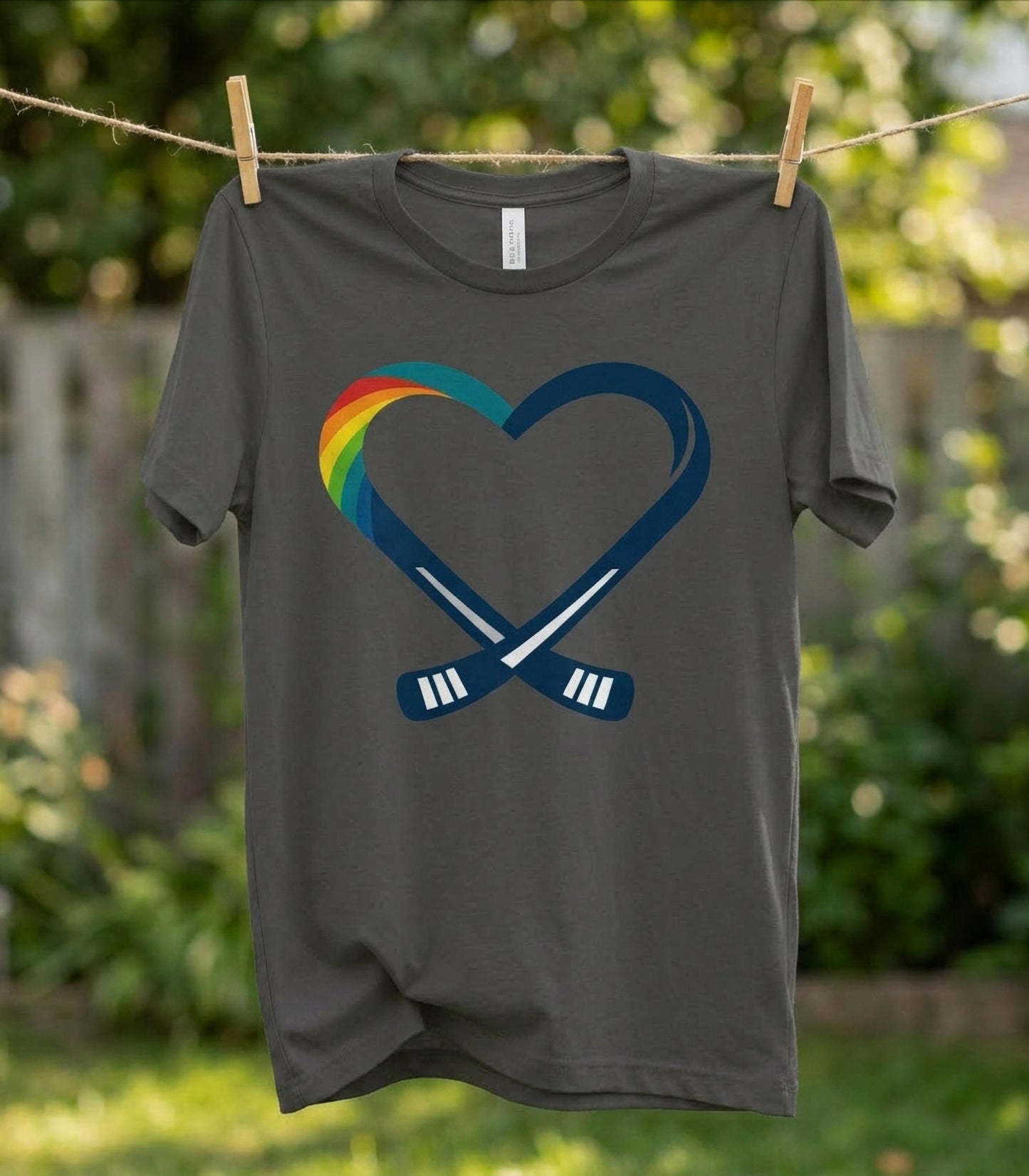 Gray rainbow heart inclusive ring spun cotton t-shirt with a heart and hockey stick design hanging on a clothesline outdoors.