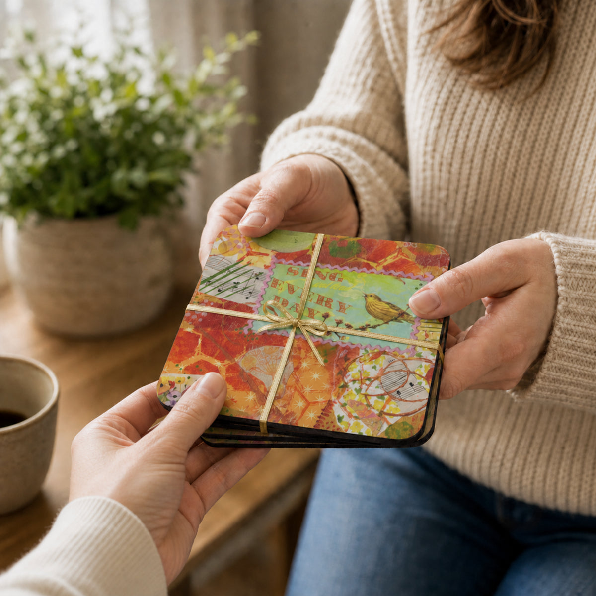 A person in a beige ribbed sweater holds a small wrapped set of art coasters, while another person's hands receive it in a softly lit indoor setting with a plant and cup in the background.