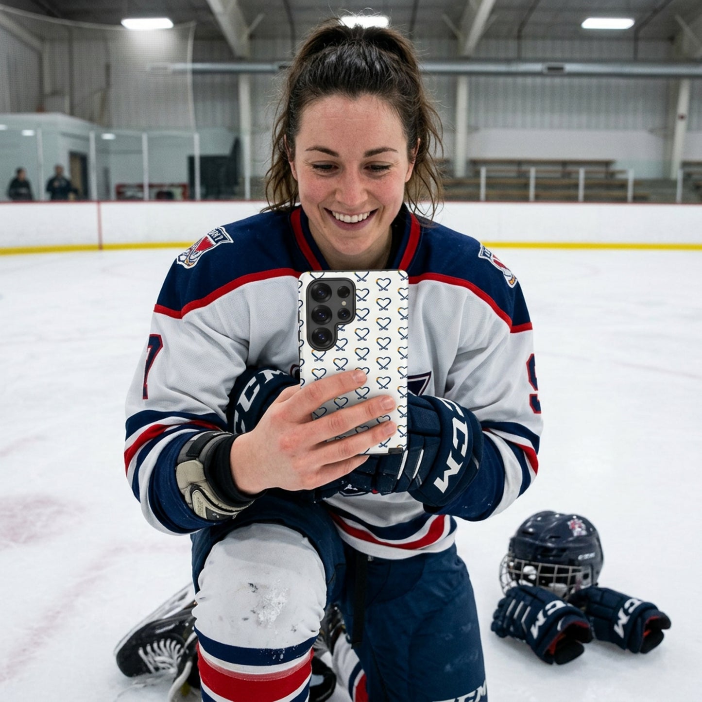Person wearing hockey gear, on her knees on an ice rink holding a Samsung phone with a rainbow heart hockey logo repeated on the phone cover, taking a selfie 