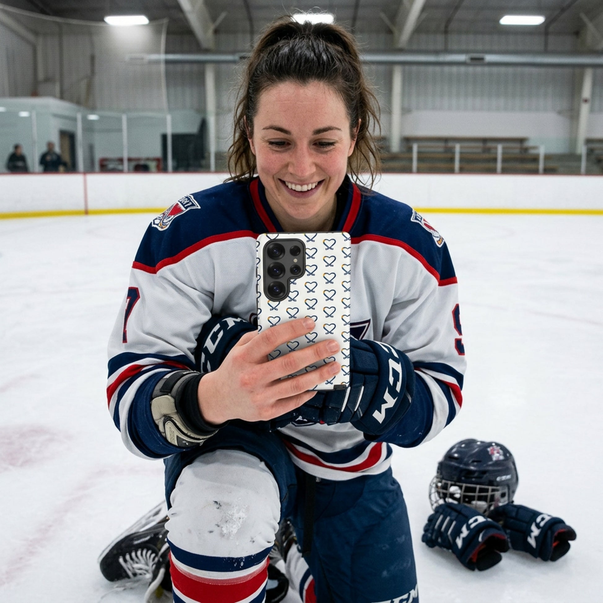 Person wearing hockey gear, on her knees on an ice rink holding a Samsung phone with a rainbow heart hockey logo repeated on the phone cover, taking a selfie 