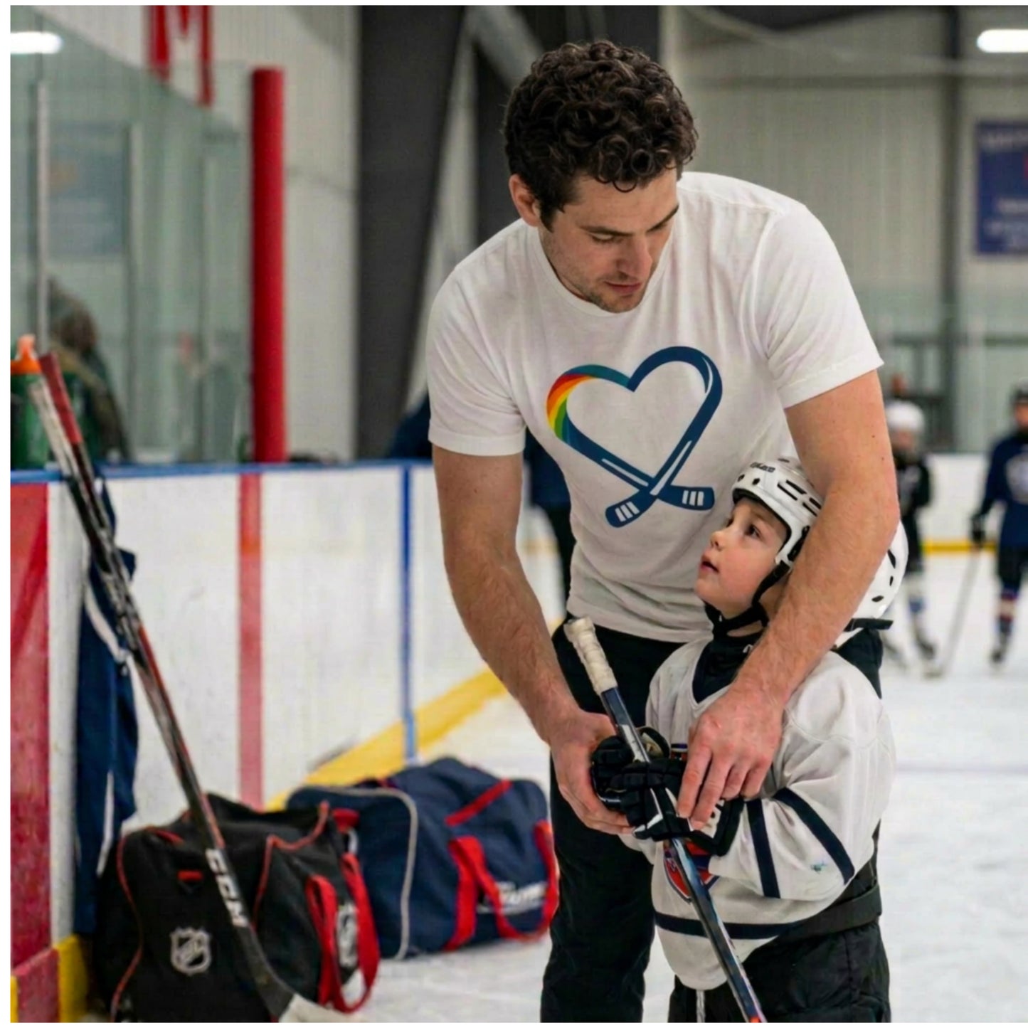 Man  is wearing the heart hockey logo t-shirt while teaching a child ice hockey on an indoor rink.