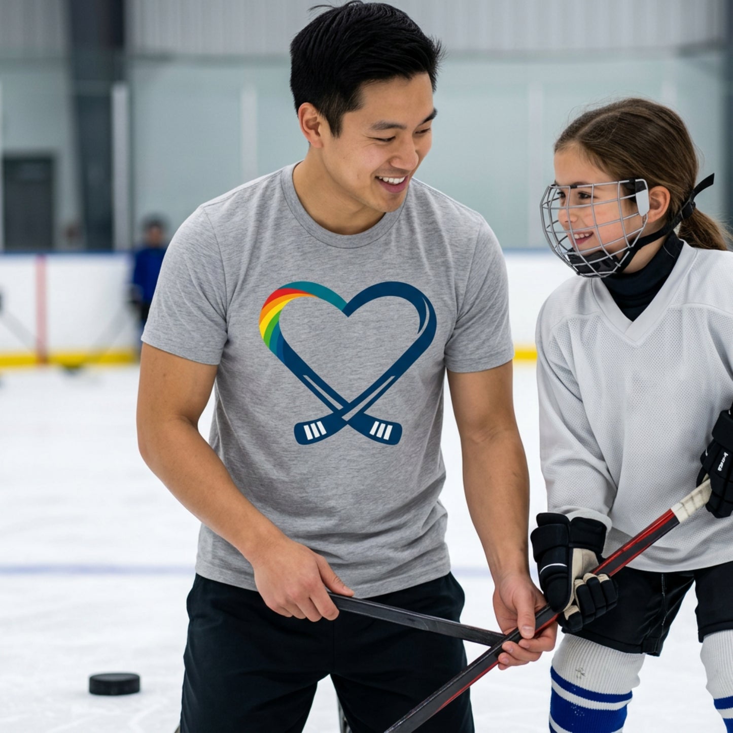 Two people on an ice rink, one wearing a gray t-shirt with a hockey heart and rainbow design, both people are holding hockey sticks.