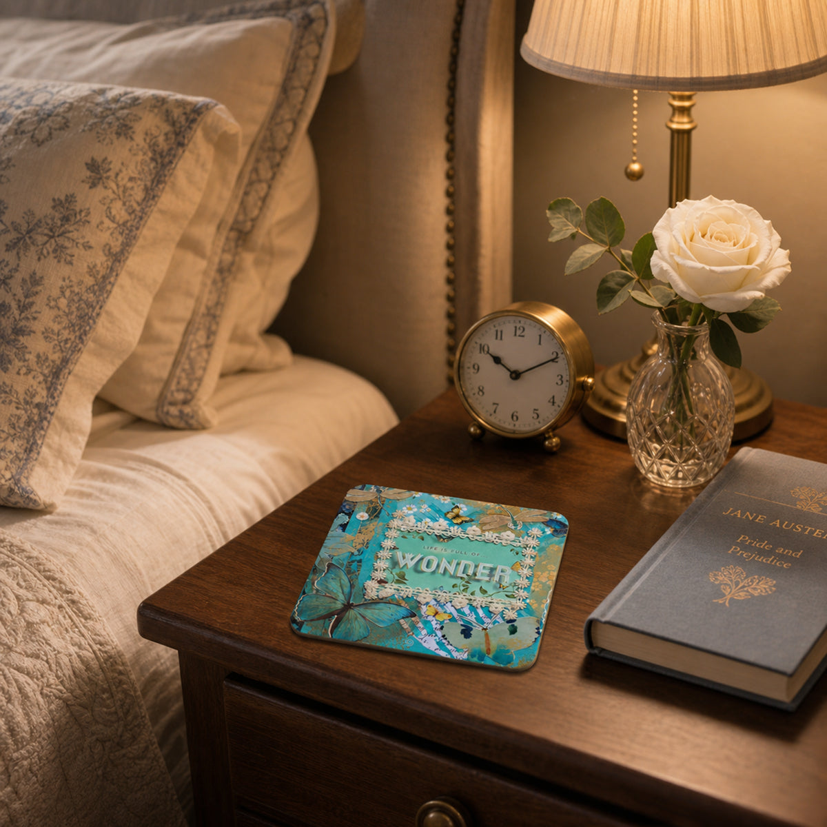 A square decorative coaster with a colorful floral and butterfly pattern and the word 'WONDER' in the center, styled on a wooden bedside table next to a lamp, a book, and a vase of rose.