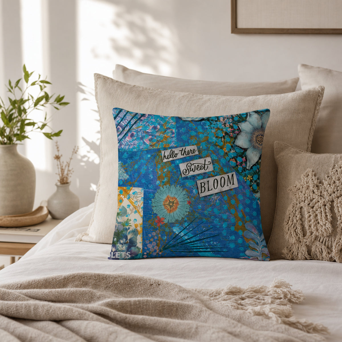 A decorative square throw pillow with a colorful floral and typographic design reading 'hello there Sweet BLOOM', styled on a bed with neutral beige bedding and pillows in a softly lit bedroom.