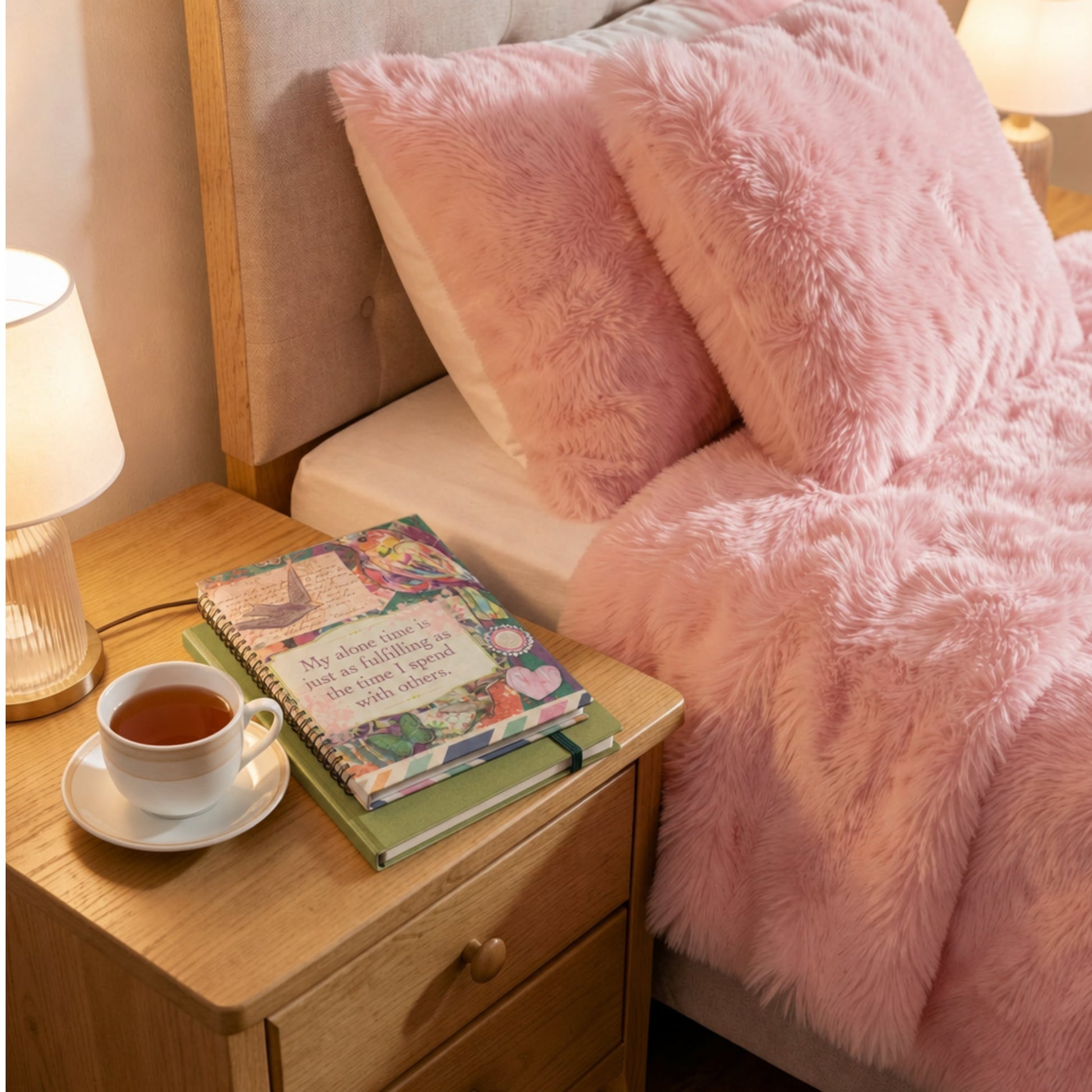 Pink fluffy blanket draped over a bed with a nightstand where the Affirmation Journal rests and a cup of tea.