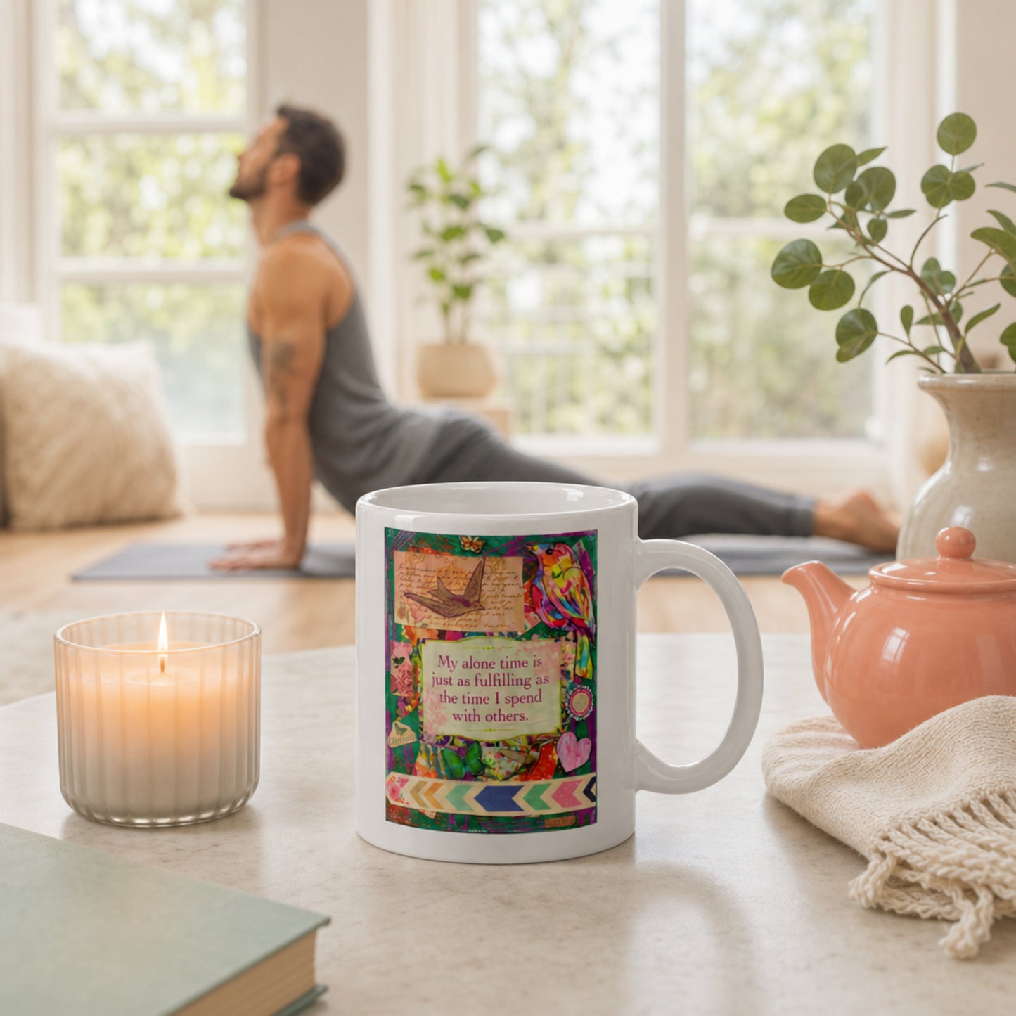 Mug with colorful design and text on a table with a candle, book, and teapot in a bright room.