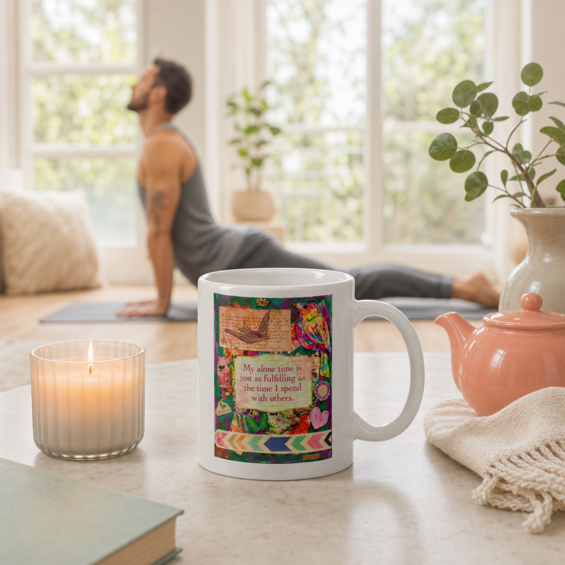 Mug with colorful design and text on a table with a candle, book, and teapot in a bright room.