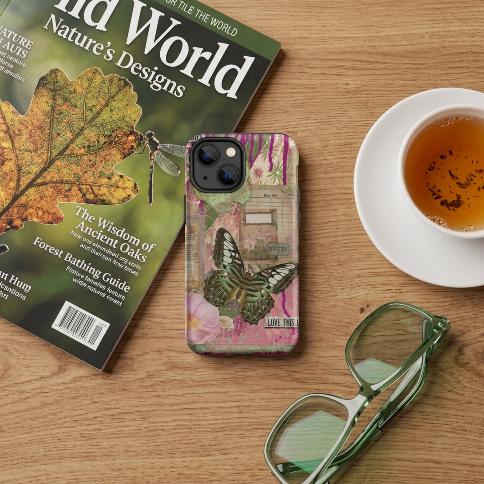 Wooden table with a magazine, cup of tea and eyeglasses, and the pink floral butterfly phone case with love this life quote next to them.