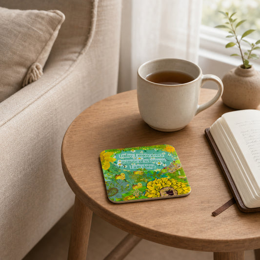 A square coaster with a colorful floral design and a quote sits on a round wooden side table in a cozy living room setting, next to a cup of tea, an open book, and a small potted plant.